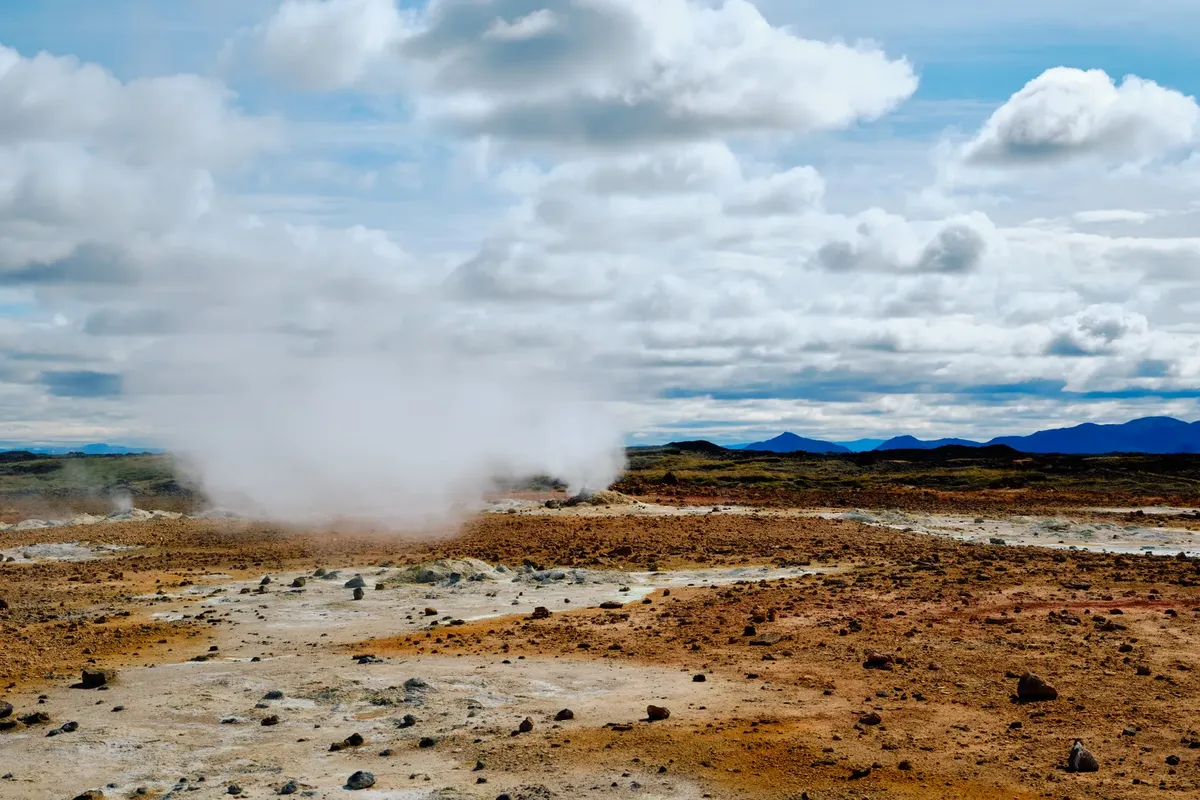 Wide view of a steaming geothermal field with cloudy sky, showcasing natural geothermal activity.